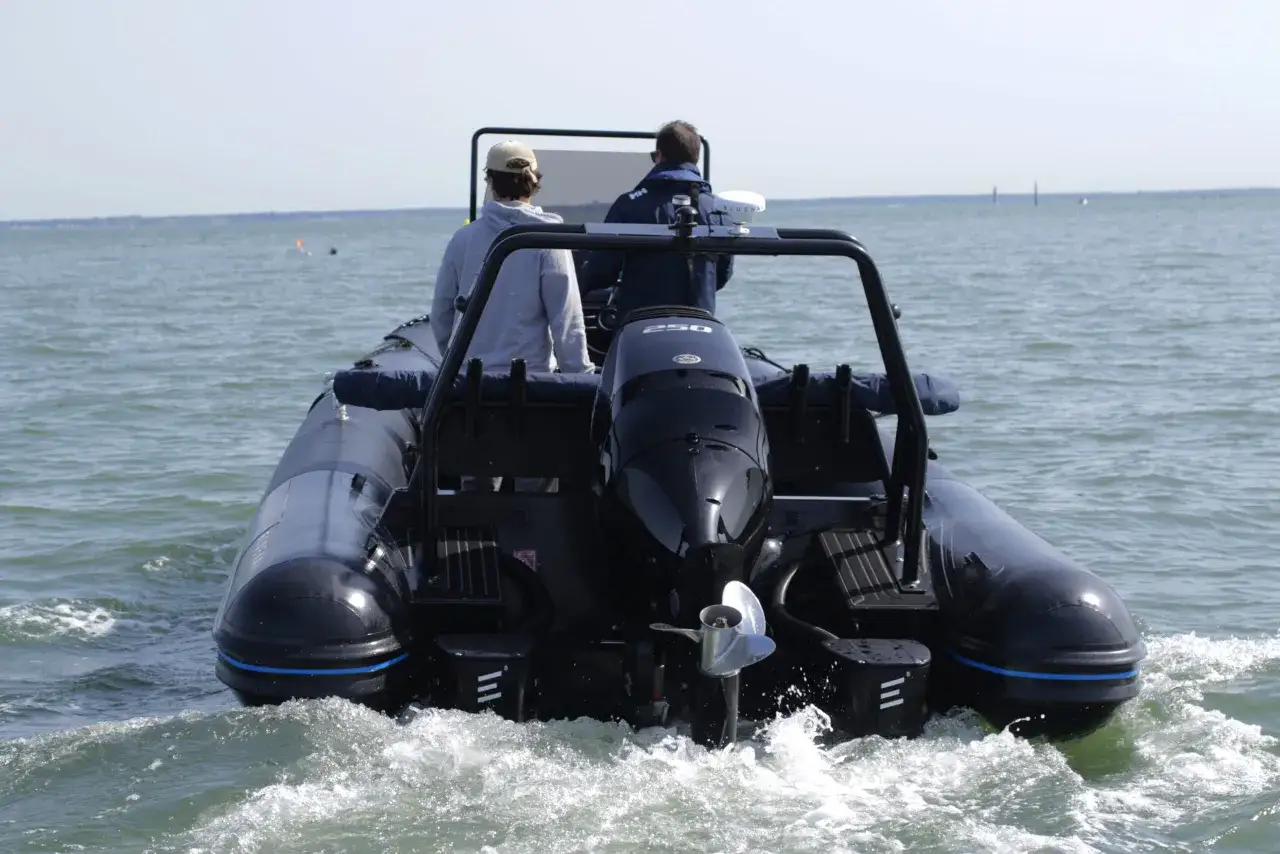 Deux personnes naviguent sur un bateau pneumatique noir, propuls&eacute; par un moteur hors-bord. L'eau &eacute;clabousse autour du bateau &eacute;lectrique.