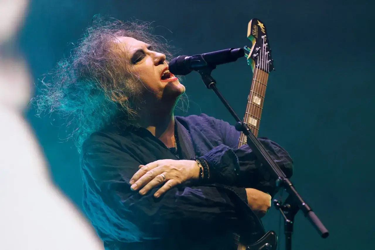 Robert Smith von The Cure singt mit geschlossenen Augen und zerzaustem Haar ins Mikrofon, w&auml;hrend er seine Gitarre h&auml;lt.