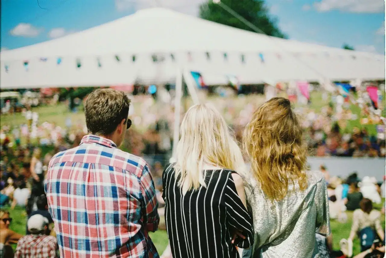 Tre vänner njuter av en solig dag på en musikfestival i Stockholm. En stor vit tältduk skymtar i bakgrunden, dekorerad med färgglada girlanger.
