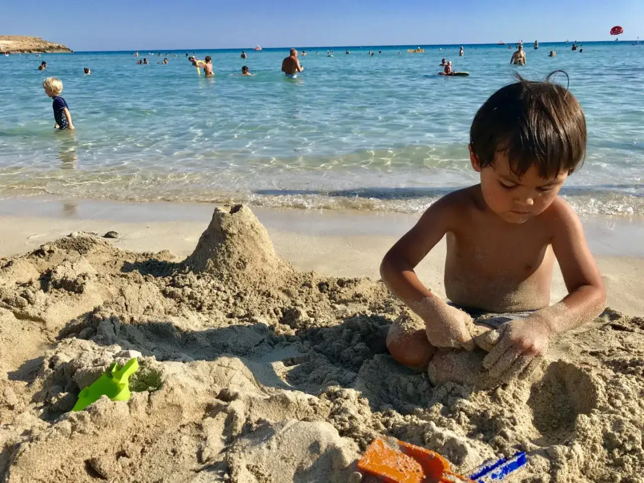 Chłopiec buduje zamek z piasku na plaży, idealne cypr atrakcje dla dzieci. W tle widać morze i innych plażowicz&oacute;w.