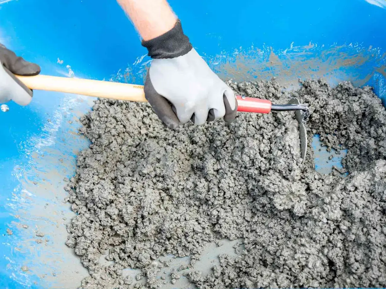 Manos enguantadas mezclan cemento y agua en una carretilla azul, aprendiendo como hacer hormig&oacute;n para suelo.