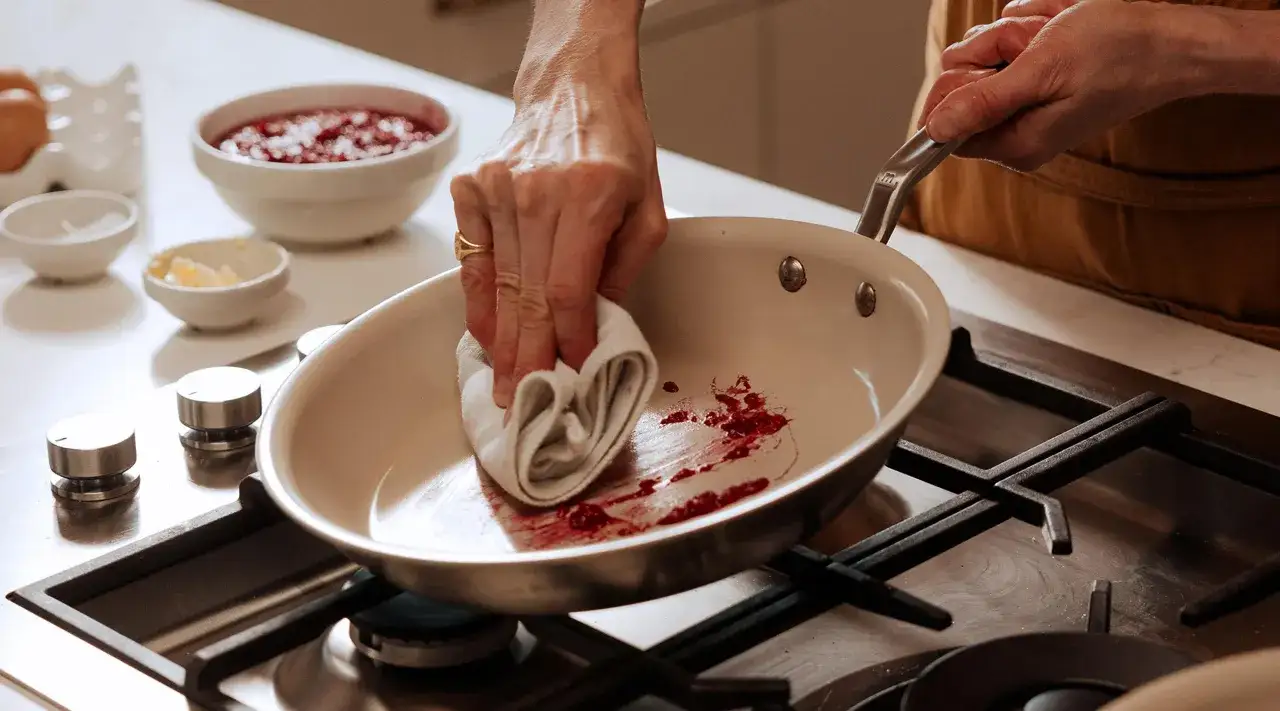 En hand torkar ur en stekpanna med en trasa, rester av röda bär syns. Ska man diska stekpannan med diskmedel?