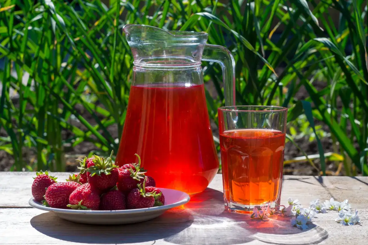 En karaff och ett glas med r&ouml;d saft st&aring;r p&aring; ett tr&auml;bord. En tallrik med f&auml;rska jordgubbar bredvid. Perfekt f&ouml;r ett enkelt jordgubbssaft recept.