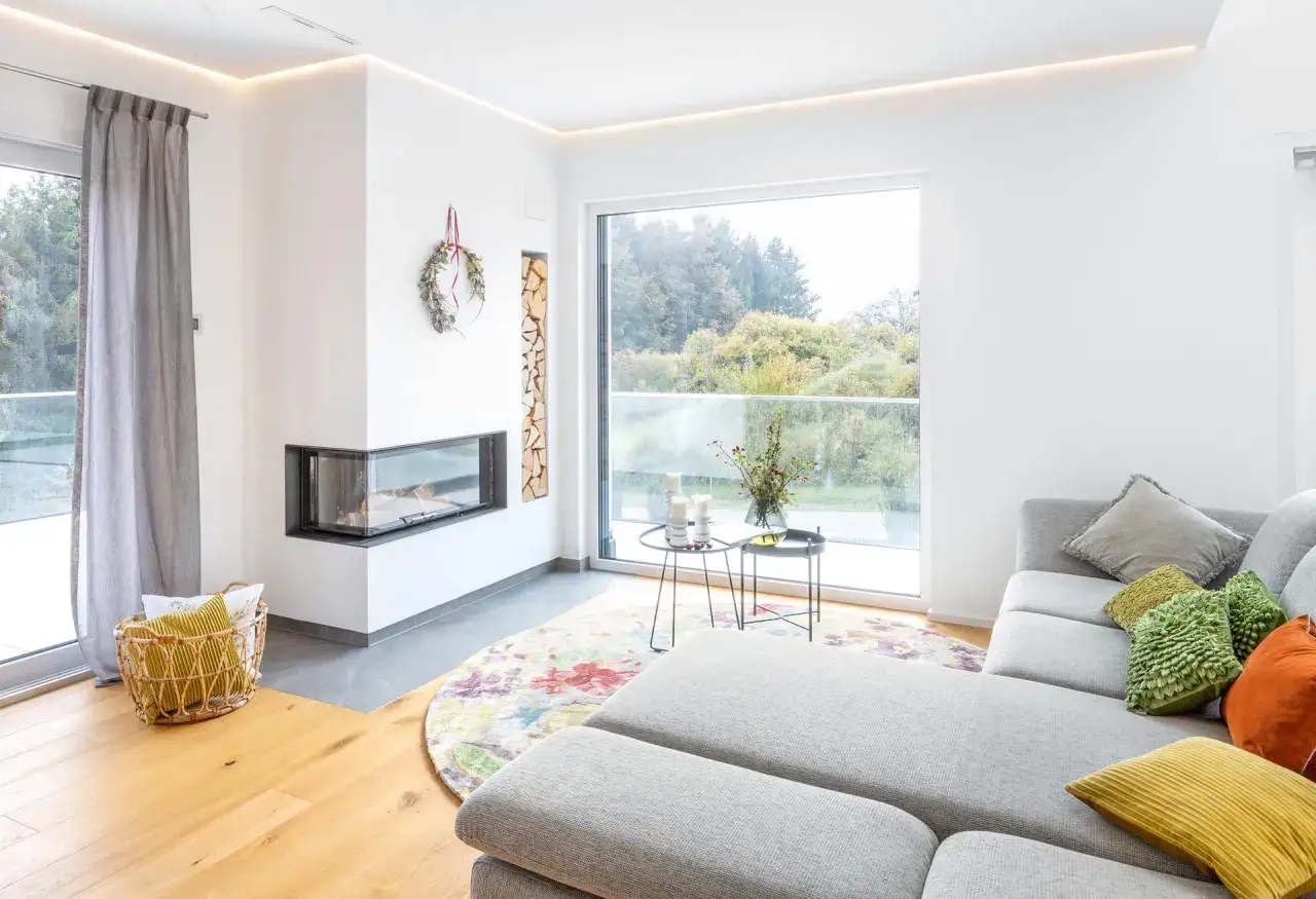 Moderner Wohnraum mit Eckkamin und Holzstapel, der den Unterschied zwischen Kamin und Ofen verdeutlicht. Gemütliche Couch mit bunten Kissen.