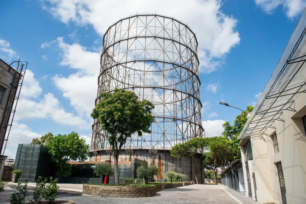 Il gasometro di Roma, una struttura imponente con un reticolo metallico, si erge sotto un cielo azzurro con nuvole bianche.