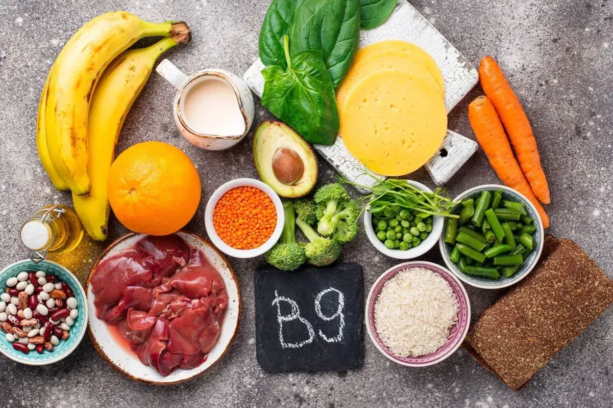 Wyszukujesz produkty bogate w witaminę B9? Na stole znajdziesz wątróbkę, szpinak, brokuły, pomarańczę, fasolę, soczewicę, marchew, groszek, awokado, ser, ryż i chleb.