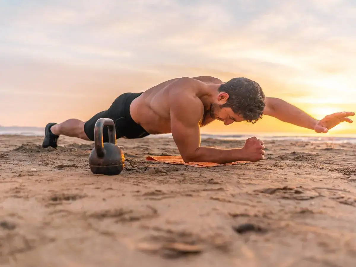 Un homme r&eacute;alise un gainage abdos sur la plage au coucher du soleil, avec une kettlebell &agrave; ses c&ocirc;t&eacute;s.
