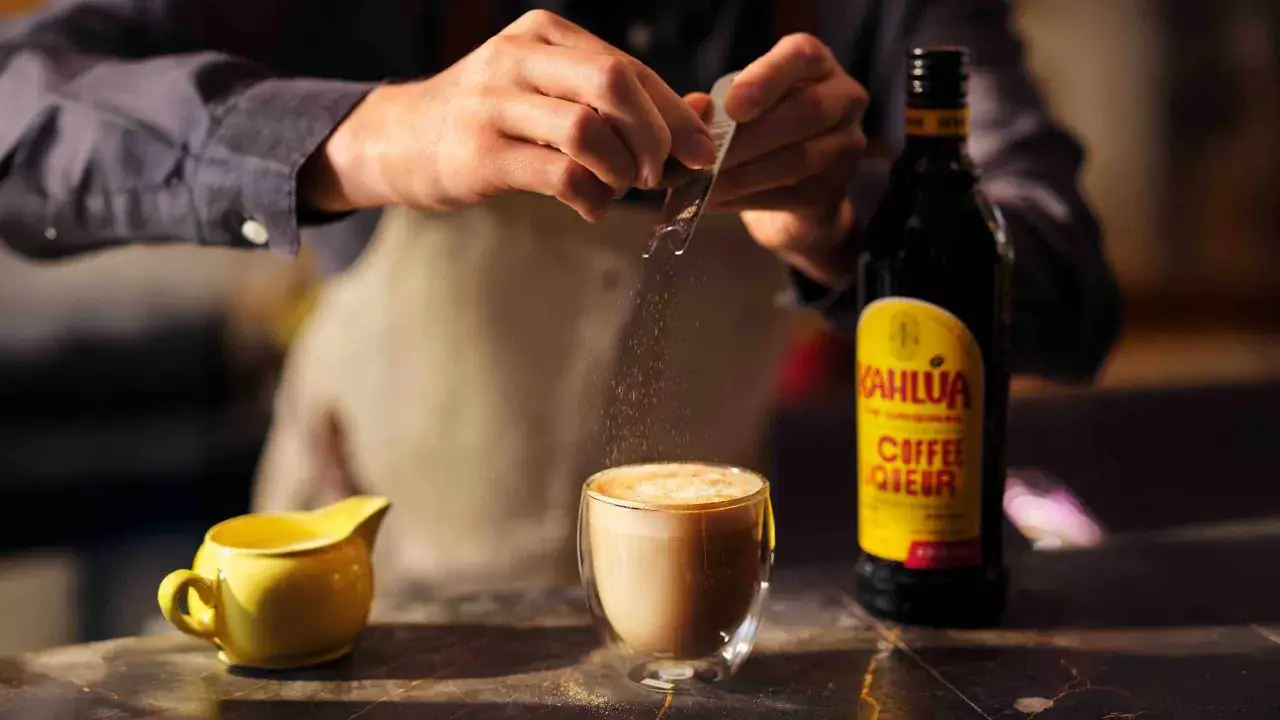 Barista dodaje cukier do kawy z Kahlúa. Butelka Kahlúa i mały dzbanuszek stoją obok.