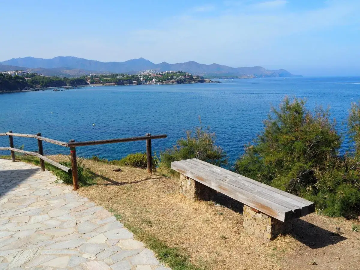 Un banco de madera espera en el camí de ronda, con vistas al azul intenso del mar y el Port de la Selva al fondo.