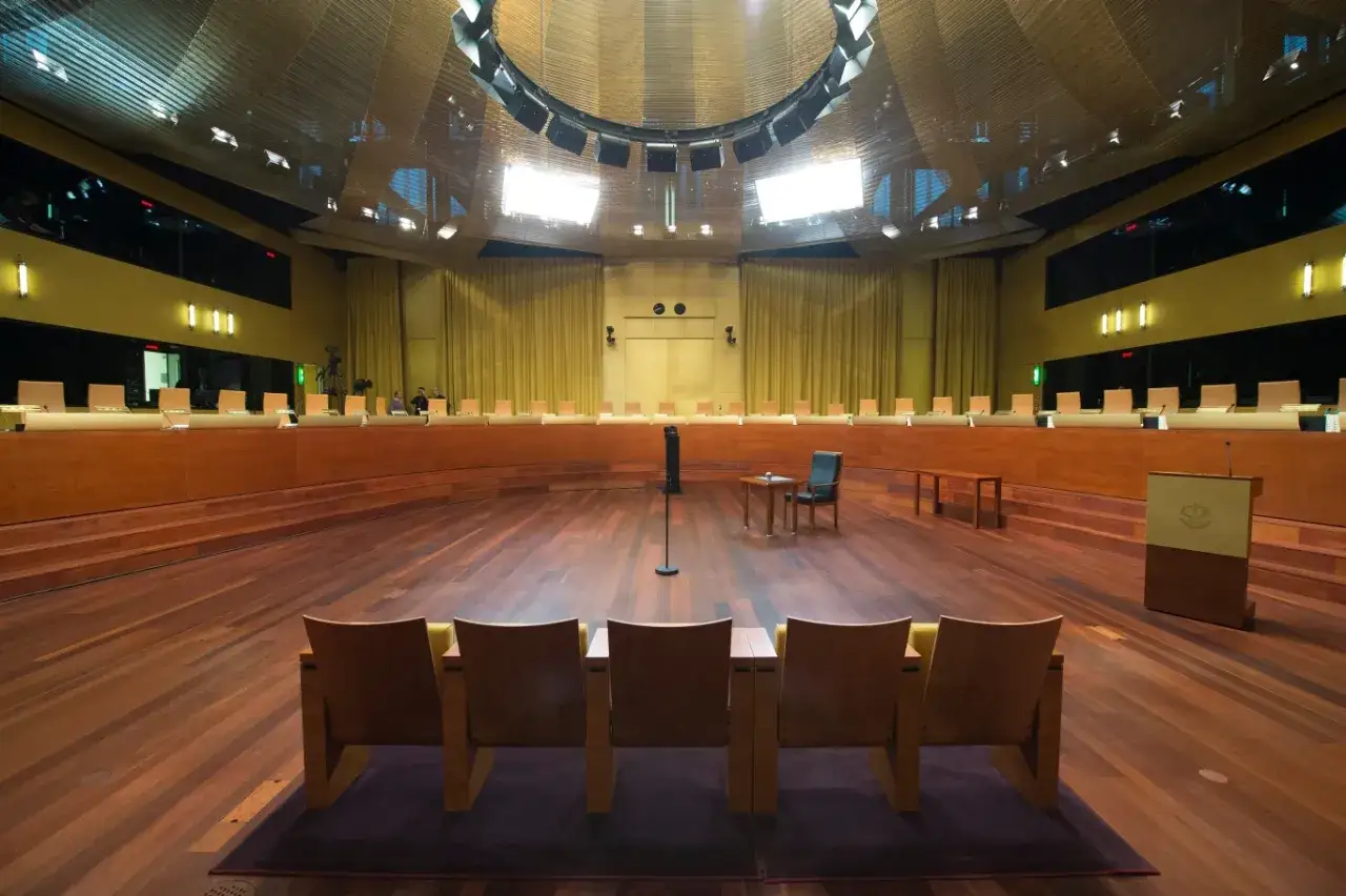 Pusta sala sądowa z drewnianą podłogą i półkolistym rzędem krzeseł. W centrum stoi mikrofon, a po prawej mównica. To miejsce, gdzie zapadają ważne **tsue**.