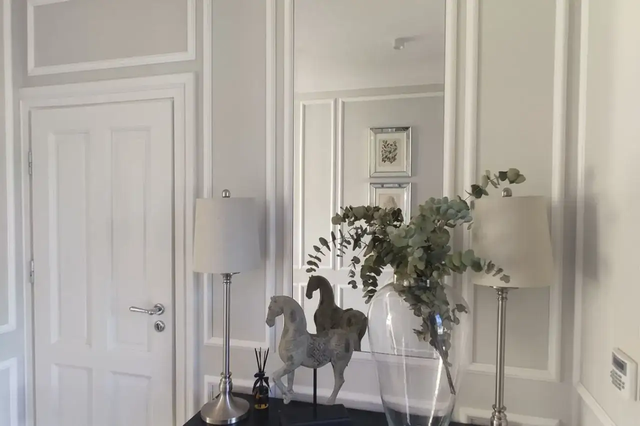 Elegancka sztukateria w przedpokoju podkreśla klasyczny styl. Dwa koniki, lampy i wazon z gałązkami dodają uroku.