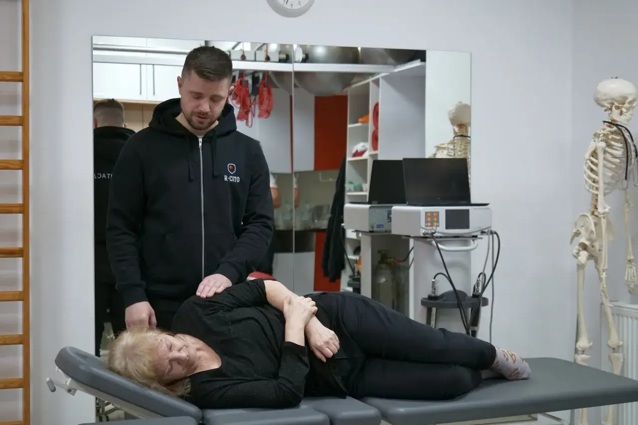 Fizjoterapeuta od kręgosłupa delikatnie dotyka pacjentki leżącej na kozetce. W tle widać szkielet i sprzęt rehabilitacyjny.