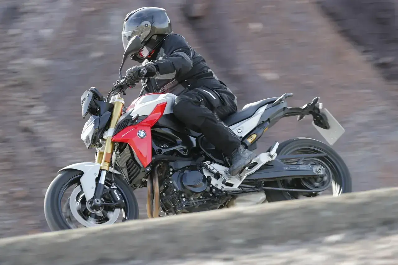 Motocyklista w kasku i kombinezonie pędzi na czerwono-białym motocyklu BMW F900R po nierównym terenie.