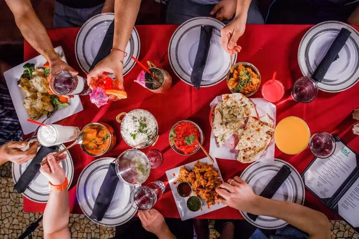 Stół zastawiony pysznym jedzeniem indyjskim: ryż, naan, curry i przekąski. Ludzie sięgają po posiłki.