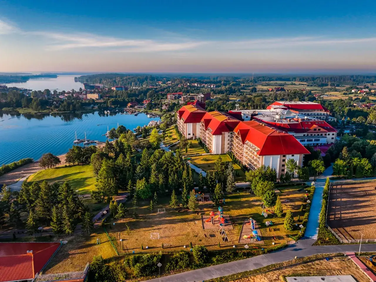 Widok z lotu ptaka na hotel Gołębiewski z czerwonym dachem, jezioro i plac zabaw. Dowiedz się, jak otrzymać złotą kartę Gołębiewski.