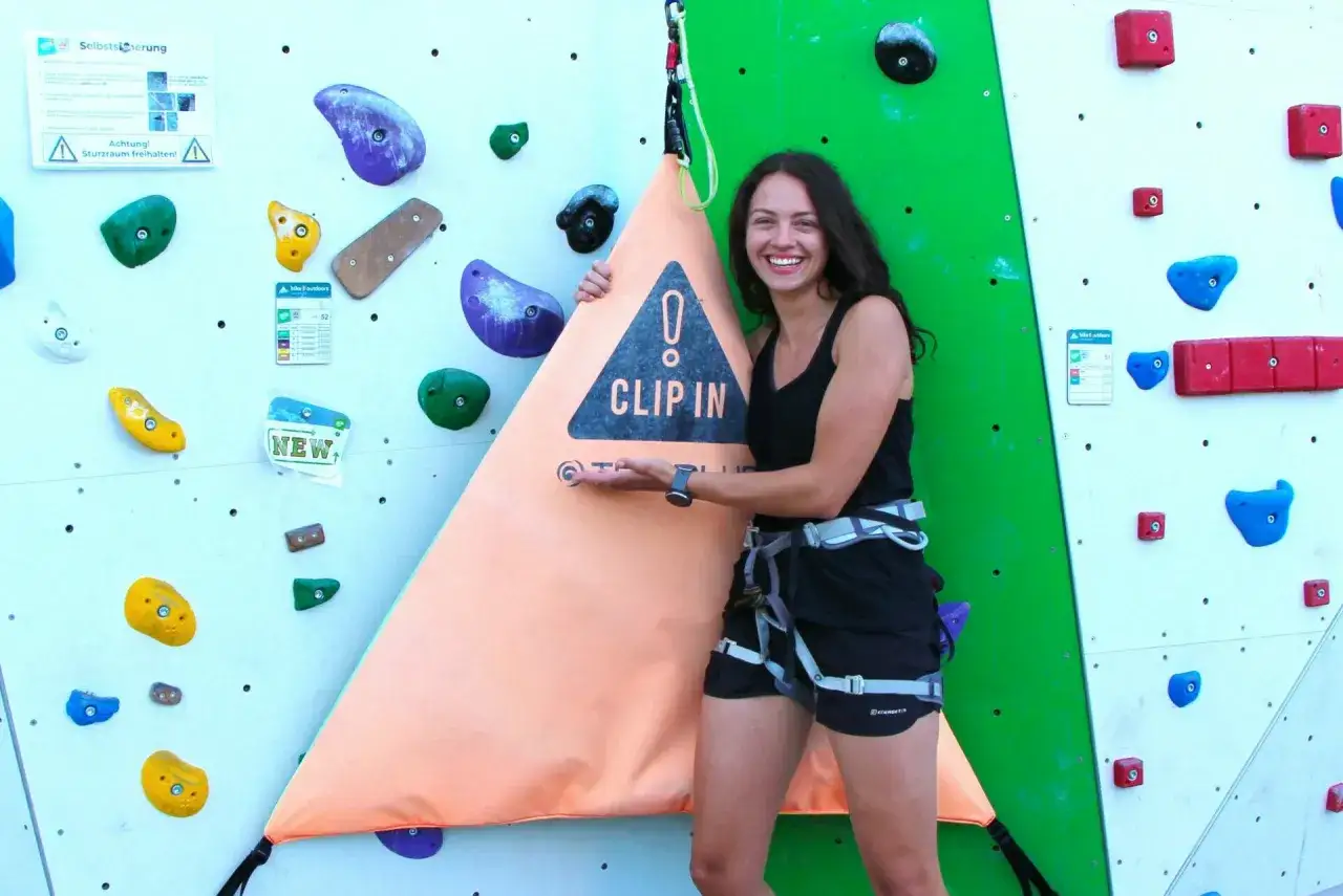 Lachende Frau mit Klettergurt vor einer Kletterwand, die auf die Wichtigkeit der selbstsicherung klettern hinweist.