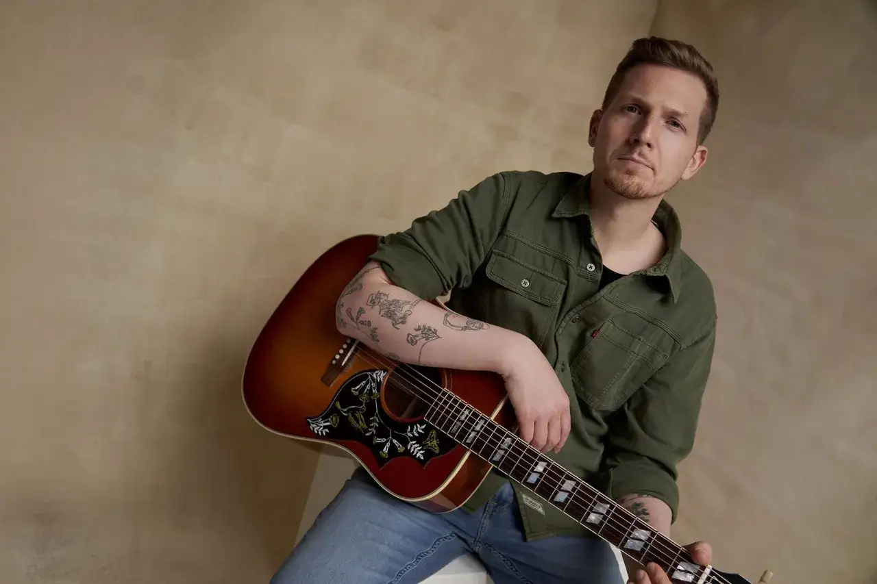 Ein Mann mit T&auml;towierungen am Arm h&auml;lt eine Gitarre. Er tr&auml;gt ein gr&uuml;nes Hemd und Jeans. Er k&ouml;nnte ein K&uuml;nstler sein, der einen Vertrag mit einem major label unterschrieben hat.