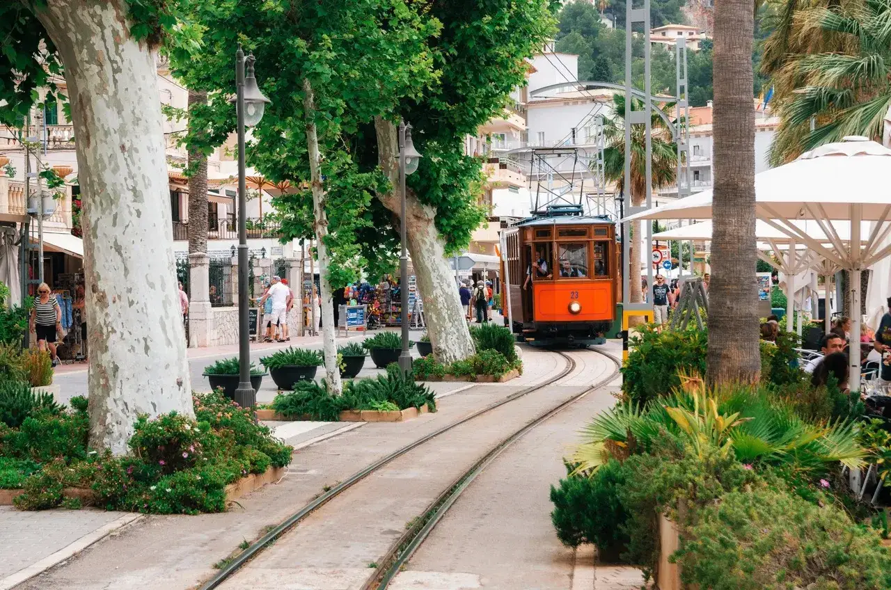En charmig orange spårvagn på väg genom en grönskande gata i Sóller, Mallorca, med vackra träd och butiker längs vägen, en perfekt vy från Palma.