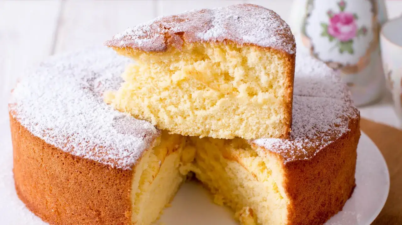 En fluffig vegansk sockerkaka pudrad med florsocker. En bit är upplagd ovanpå kakan.