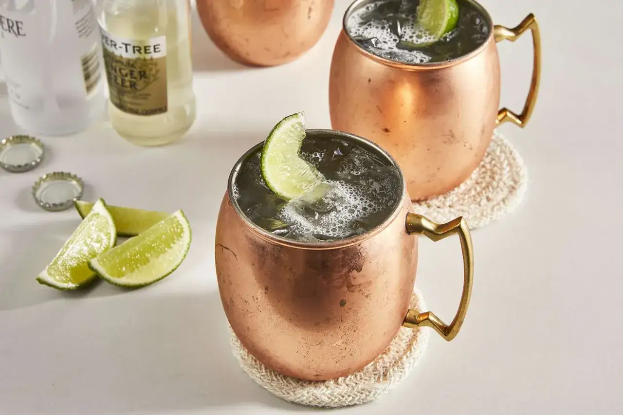 Tv&aring; koppar Moscow Mule med lime och is, redo att avnjutas. Ingredienser f&ouml;r en perfekt drink syns i bakgrunden.