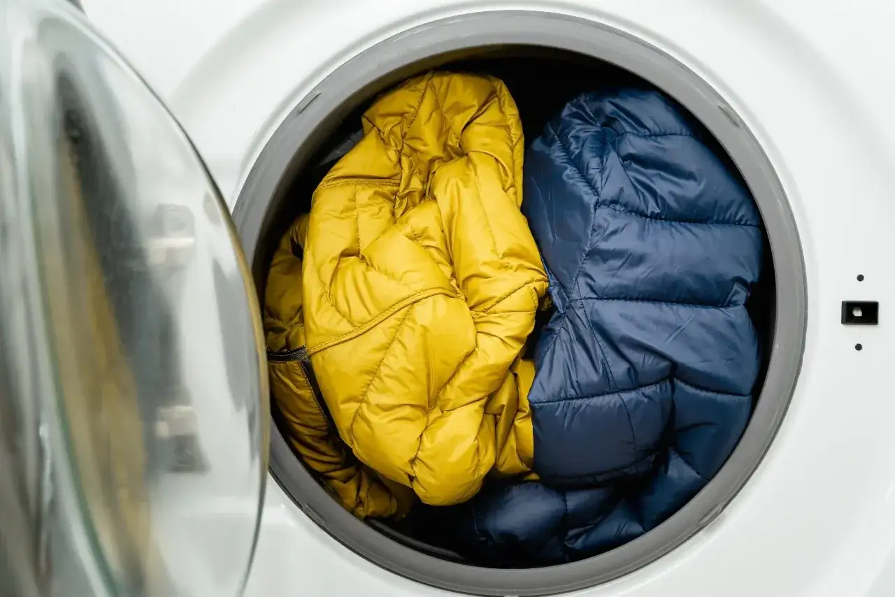 Zwei Daunenjacken, eine gelbe und eine blaue, liegen in der Trommel einer Waschmaschine. Die gelbe Jacke ist auf links gedreht, bereit f&uuml;r die W&auml;sche.