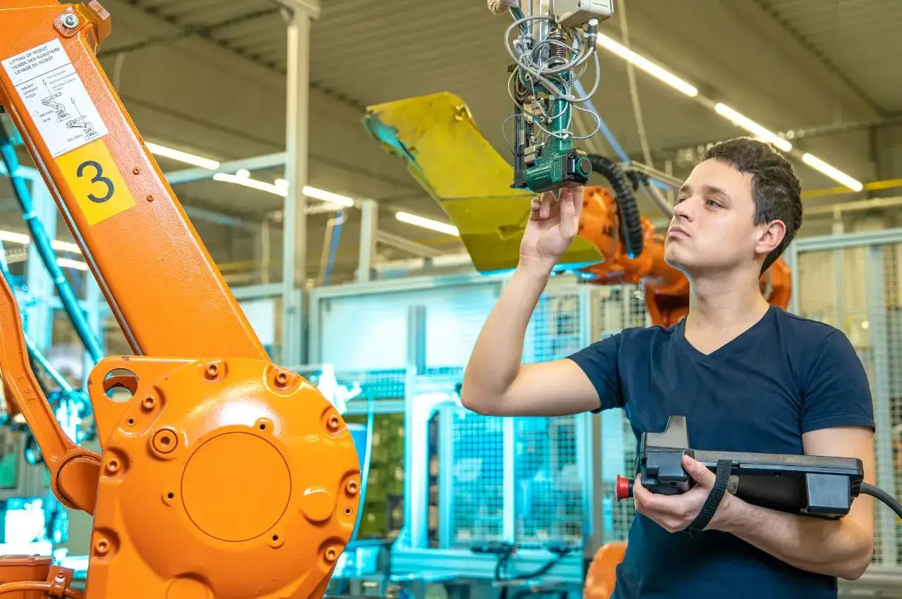 Młody inżynier z pilotem w ręku pracuje przy robocie przemysłowym. To jego praca po automatyce i robotyce.