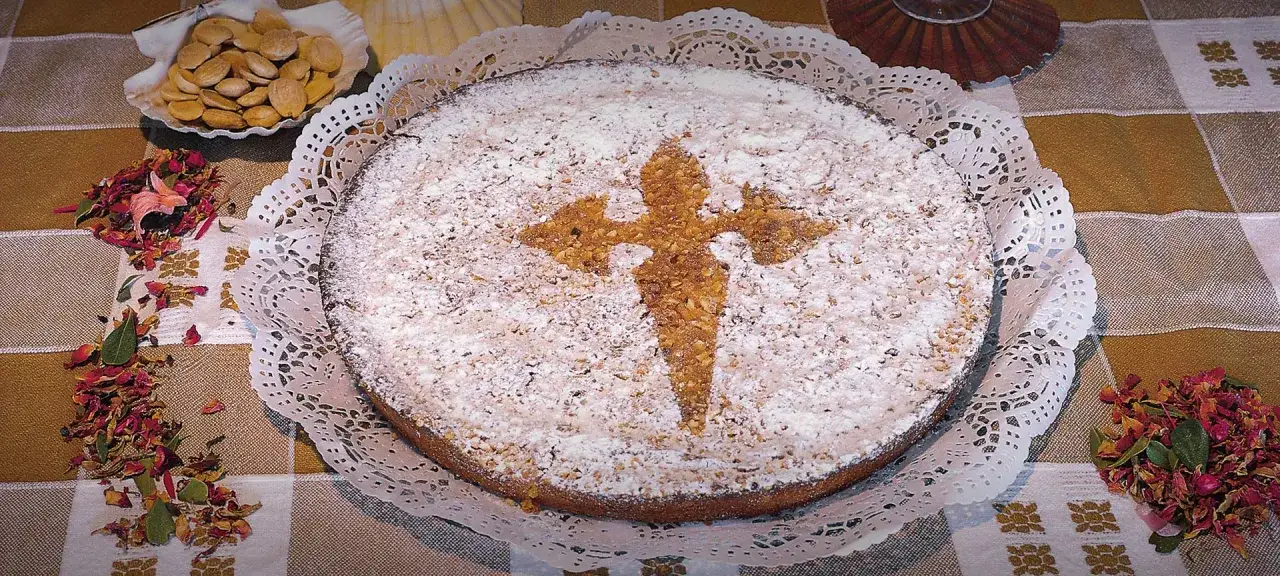 Una torta di Santiago cosparsa di zucchero a velo, con il simbolo della croce di Santiago in noci. Accanto, mandorle e petali di rosa secchi.
