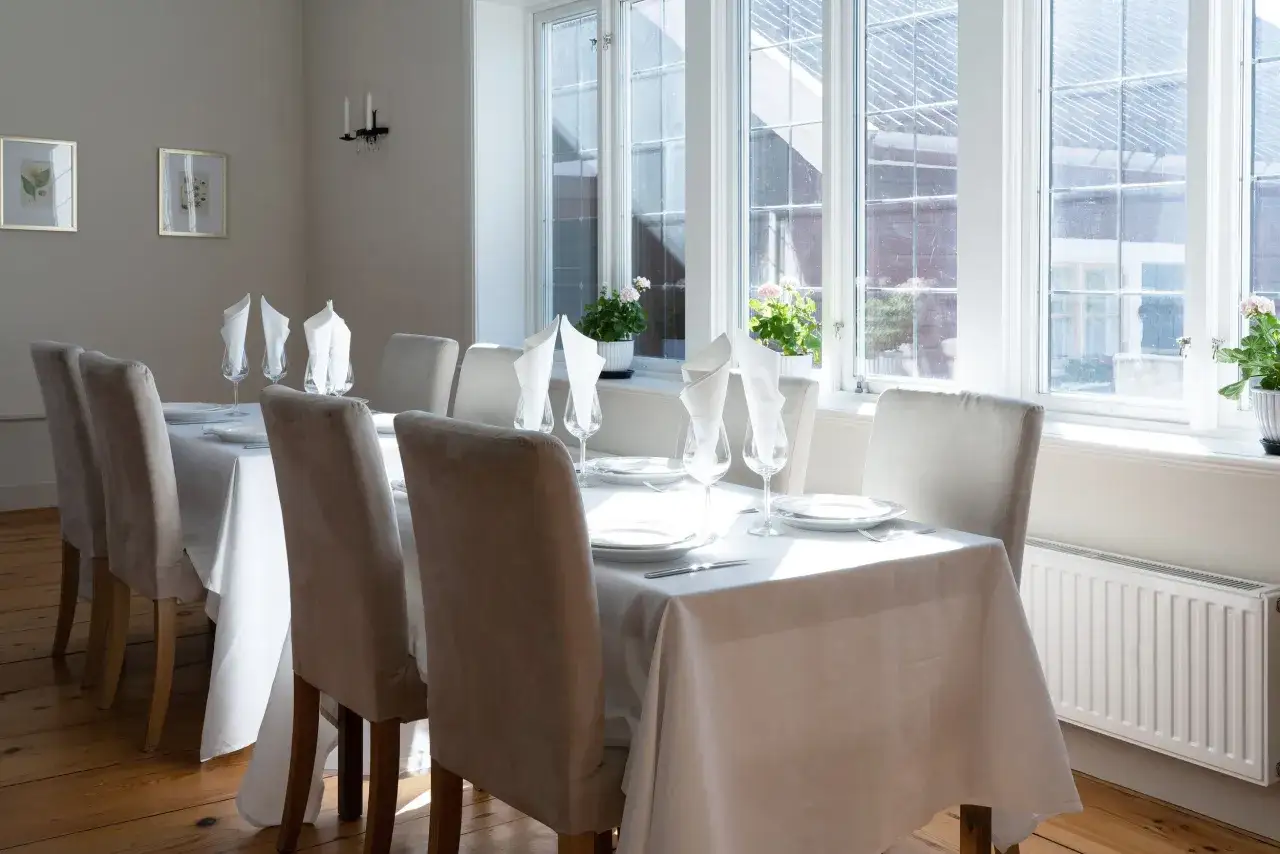Vackert dukat bord med vita linneservetter och glas i Jöns Andersgården. Solen lyser in genom fönstren.