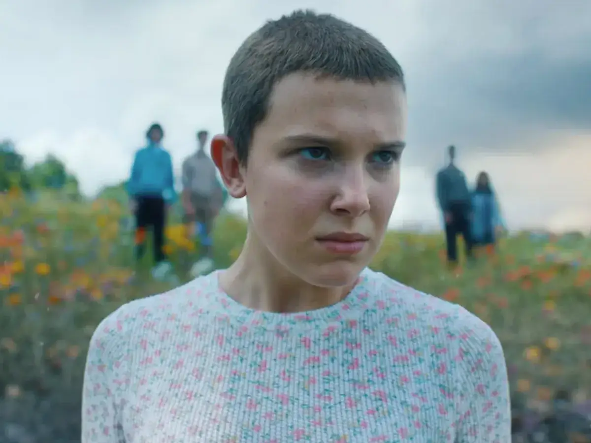 Nastka z Stranger Things w kwiecistym sweterku, z przyjaciółmi w tle, na tle pola kwiatów.