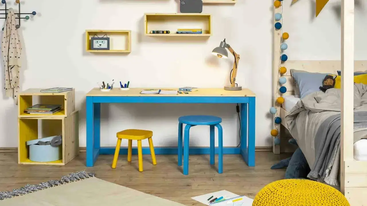 Ein **kinderschreibtisch selber bauen** ist eine tolle Idee! Hier siehst du einen hellen, modernen Schreibtisch mit blauen Beinen, daneben zwei bunte Hocker und ein Bett mit Lichterkette.