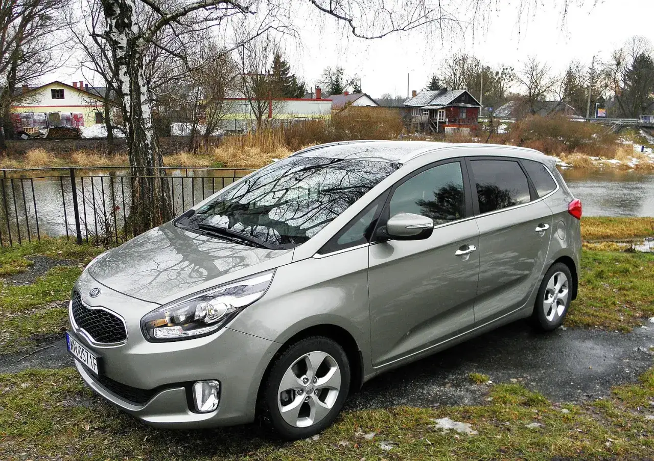 Srebrny Kia Carens 4 zaparkowany nad rzeką, z odbiciem drzew w szybie.