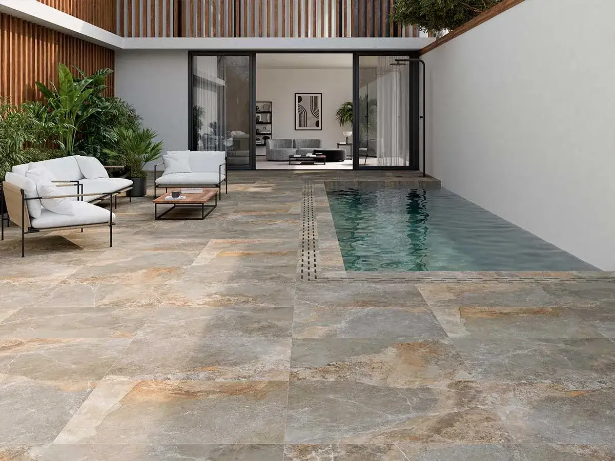 Elegante suelo para piscinas con acabado de piedra natural, rodeando una piscina de agua cristalina. Muebles de exterior y vegetación completan la escena.