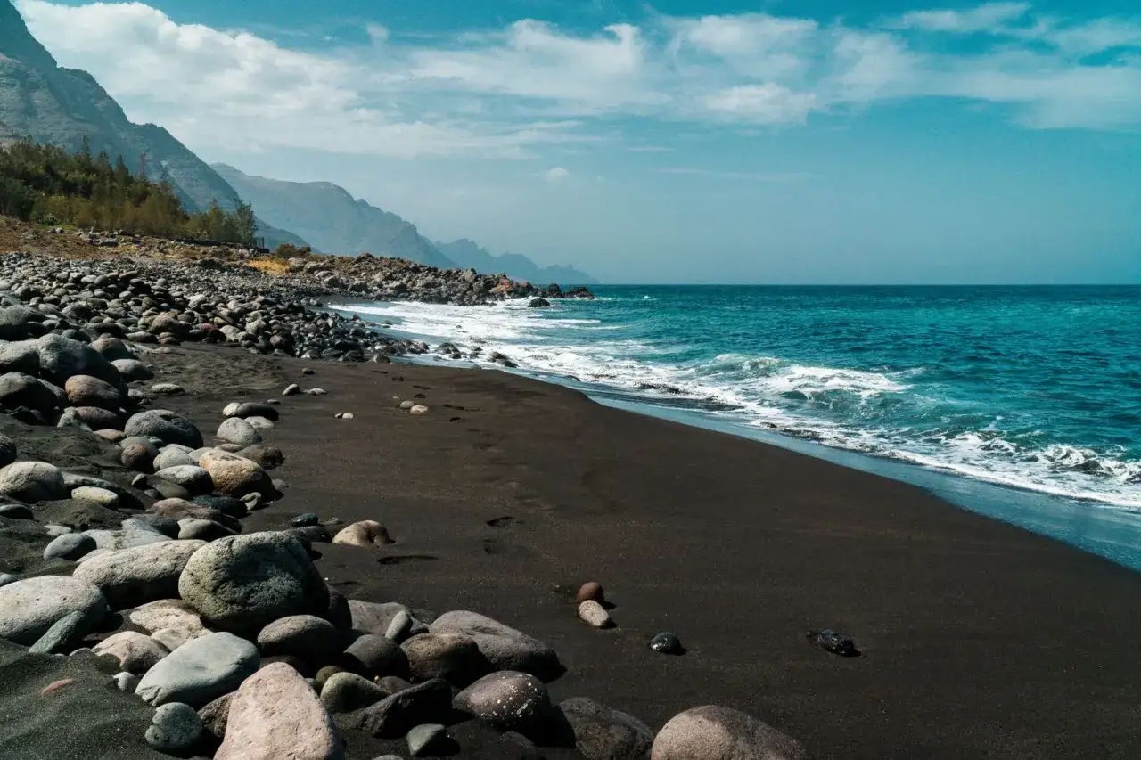 Gran Canaria schwarzer Strand mit Felsen und t&uuml;rkisfarbenem Meer unter blauem Himmel mit Wolken.