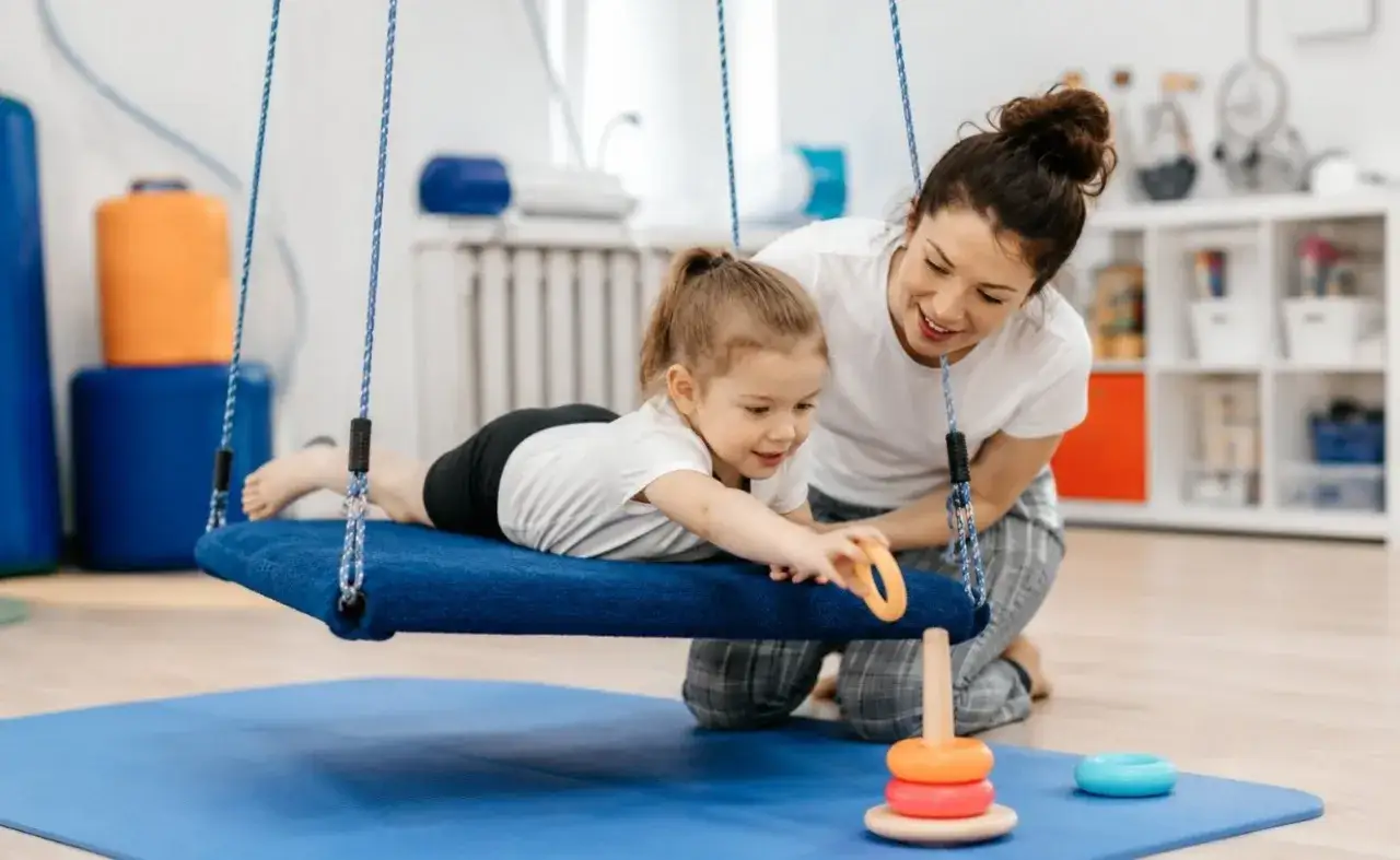 Terapia integracji sensorycznej: dziewczynka na huśtawce z terapeutką, która pomaga jej układać pierścienie.