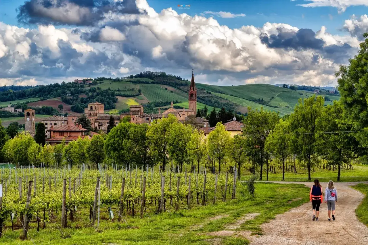 Due escursioniste passeggiano su un sentiero tra vigneti rigogliosi, con un borgo pittoresco e le dolci colline modenesi sullo sfondo.