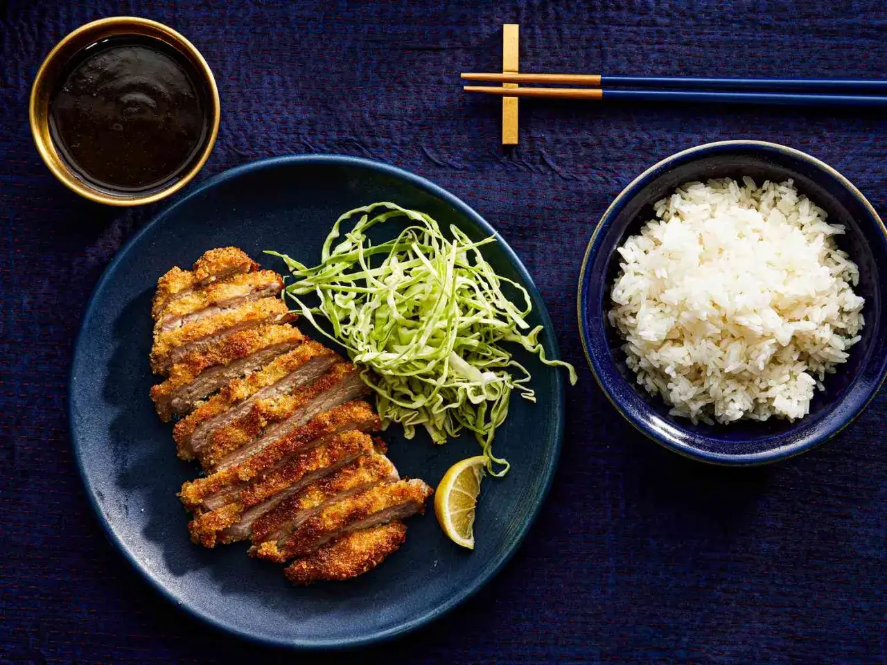 A delicious plate of baked tonkatsu, sliced and served with shredded cabbage, a lemon wedge, a bowl of rice, and a dipping sauce.