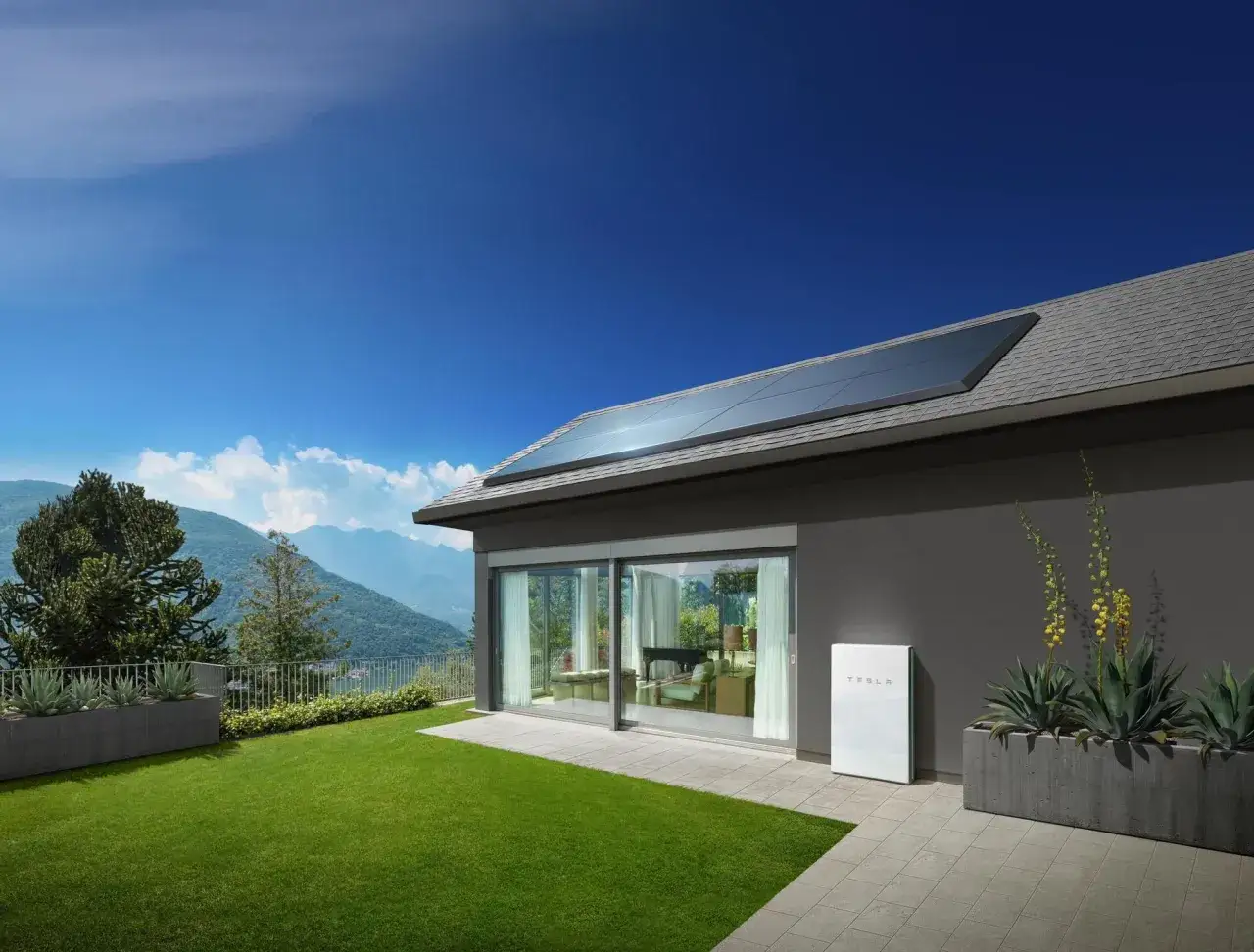Casa moderna con paneles solares en el tejado y una **tesla battery** blanca junto a plantas en el jard&iacute;n, con vistas a monta&ntilde;as.