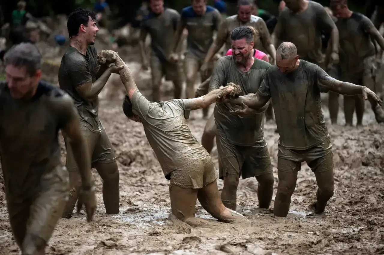 Teilnehmer kämpfen sich durch Schlamm bei einem **obstacle run**. Einer hilft dem anderen, aus dem tiefen Morast zu kommen.