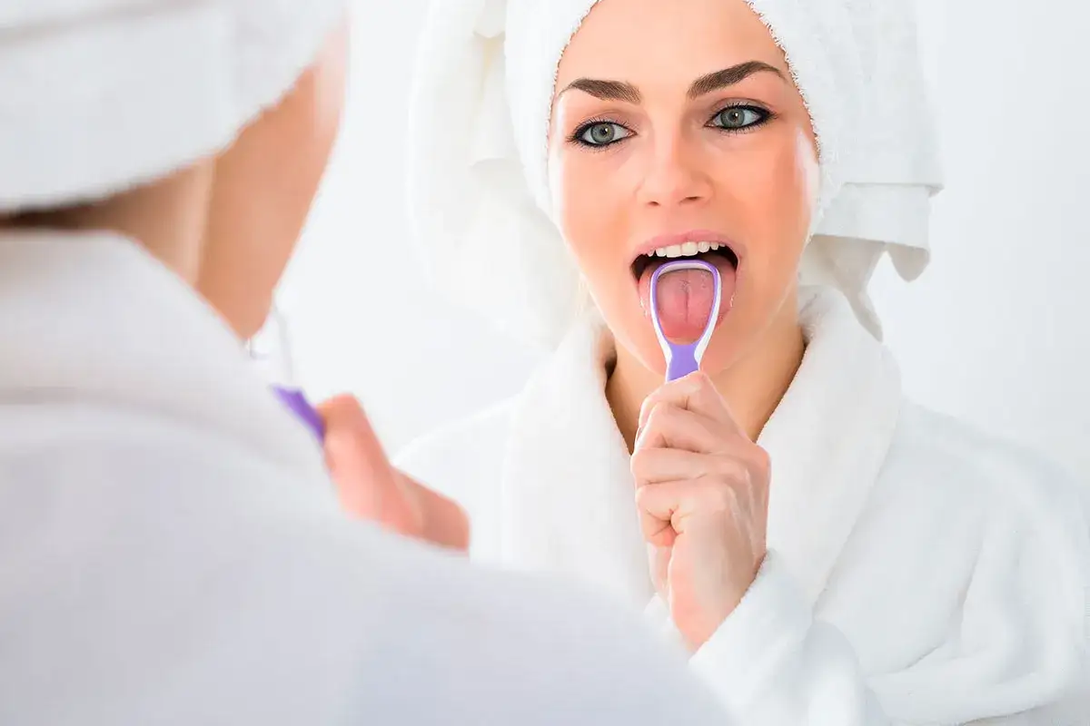 Mujer en bata y toalla se cepilla la lengua con un limpiador morado, reflejada en el espejo.