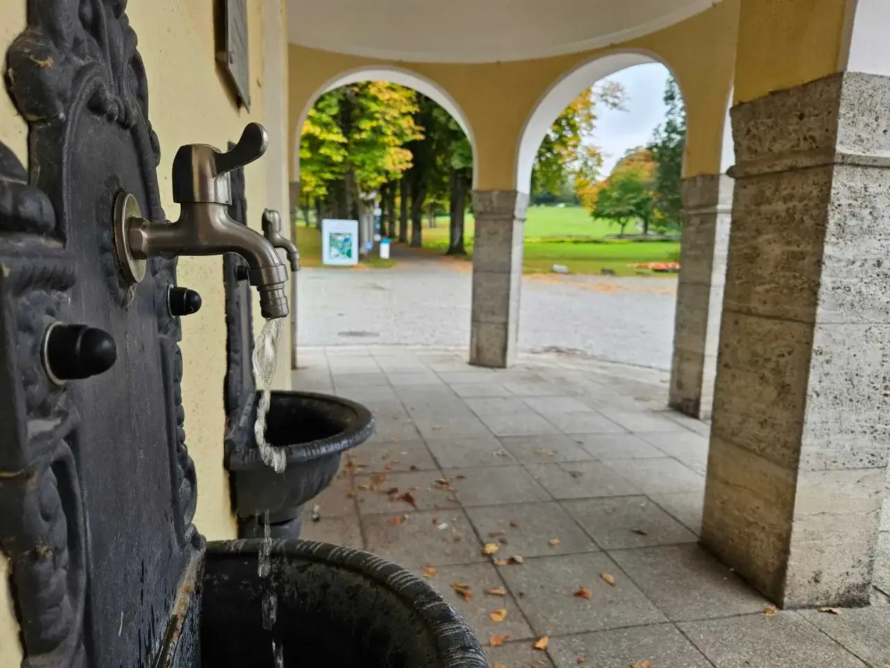 Erfrischendes Wasser flie&szlig;t aus einem historischen Brunnen im Kurpark Bad Hermannsborn. Herbstlaub liegt auf dem gepflasterten Weg.