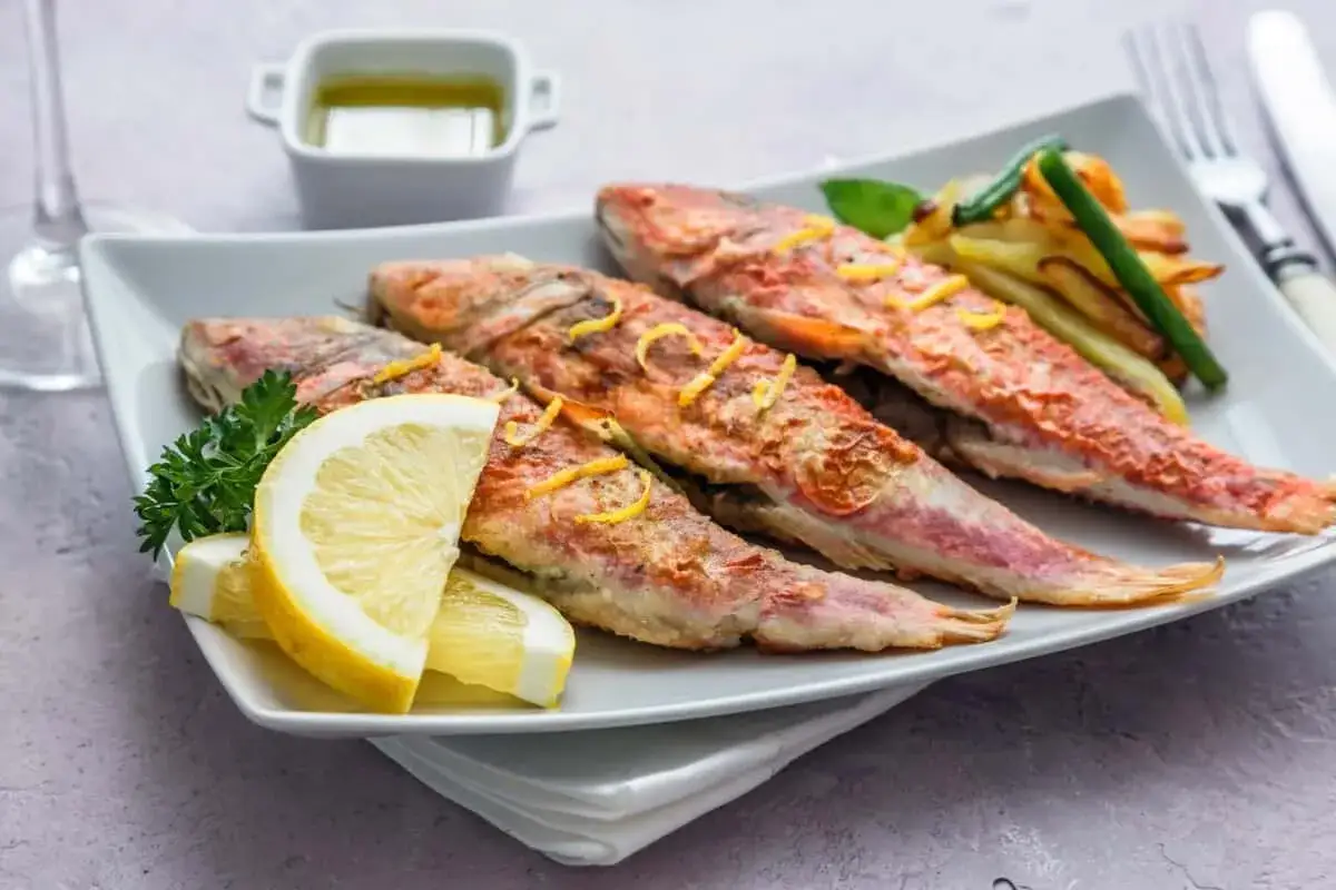 Cuatro salmonetes al horno, dorados y jugosos, servidos con rodajas de lim&oacute;n, perejil y patatas fritas.