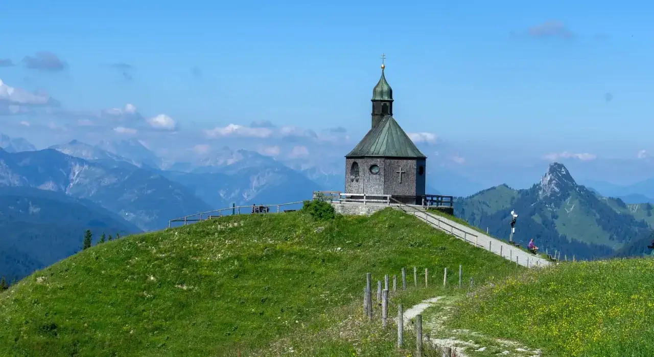 Altes Wallberghaus Wanderung: Kleine Kapelle auf einem gr&uuml;nen H&uuml;gel mit Bergpanorama. Ein Weg f&uuml;hrt zur Kapelle, die Wanderer zur Rast einl&auml;dt.