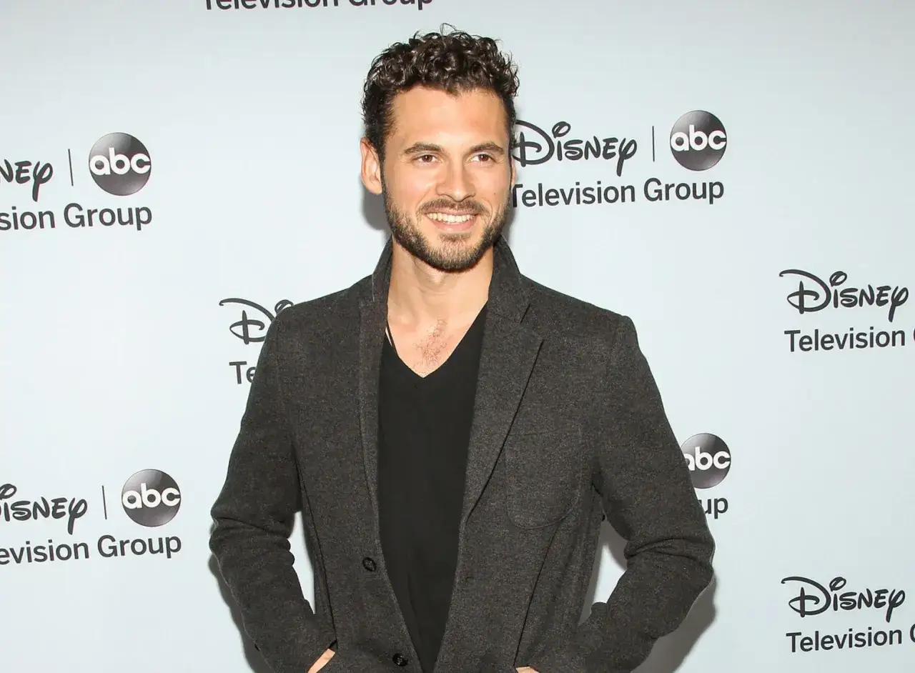 Adan Canto, med lockigt hår och skägg, ler mot kameran i en mörk kavaj över en svart tröja. Bakgrunden har Disney Television Group-logotyper.