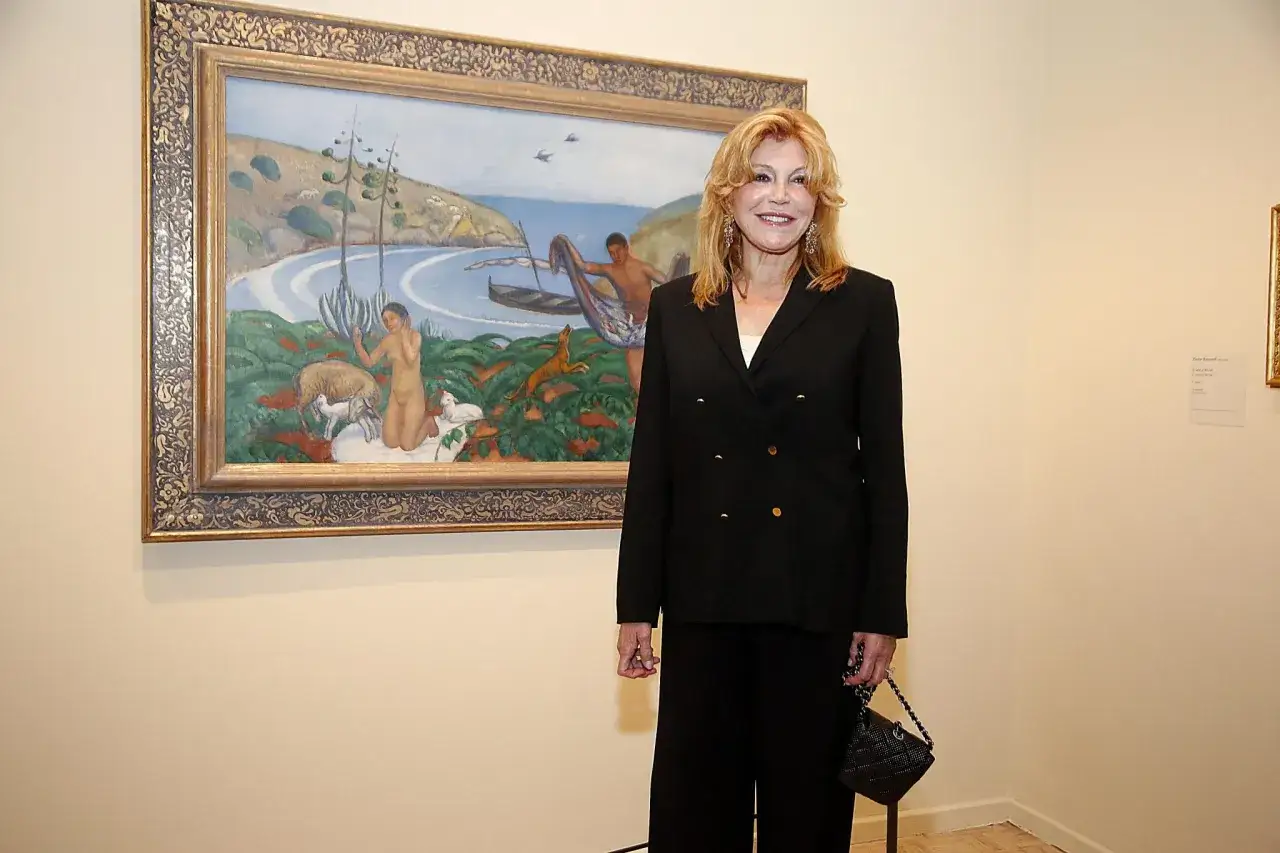 Tita Thyssen posa junto a un cuadro que representa una escena costera con figuras humanas y animales.