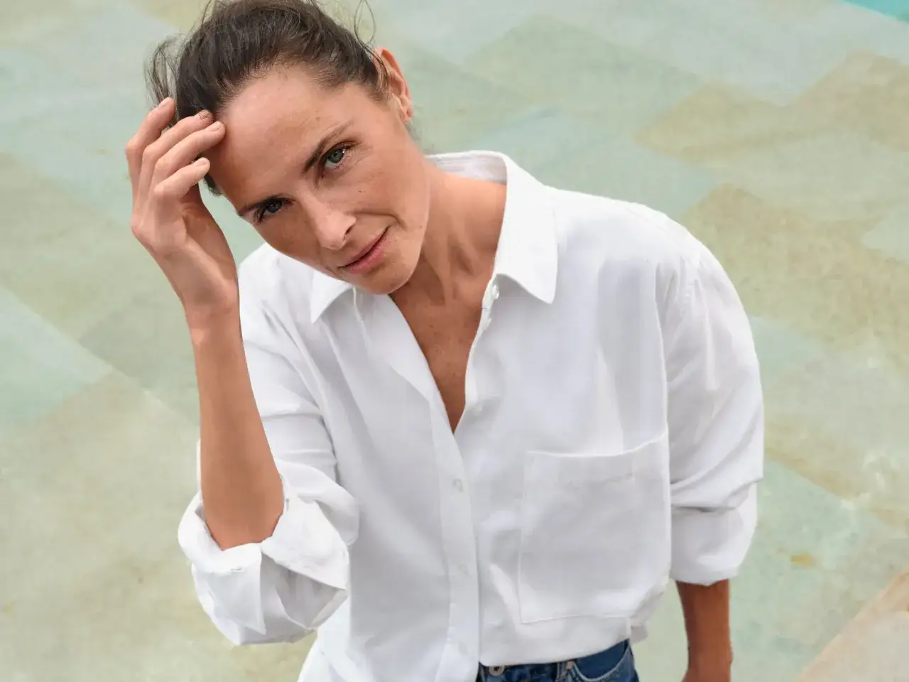 Eine Frau mit 50, die selbstbewusst in die Kamera blickt, ihre Hand leicht am Kopf. Sie tr&auml;gt ein wei&szlig;es Hemd und Jeans.