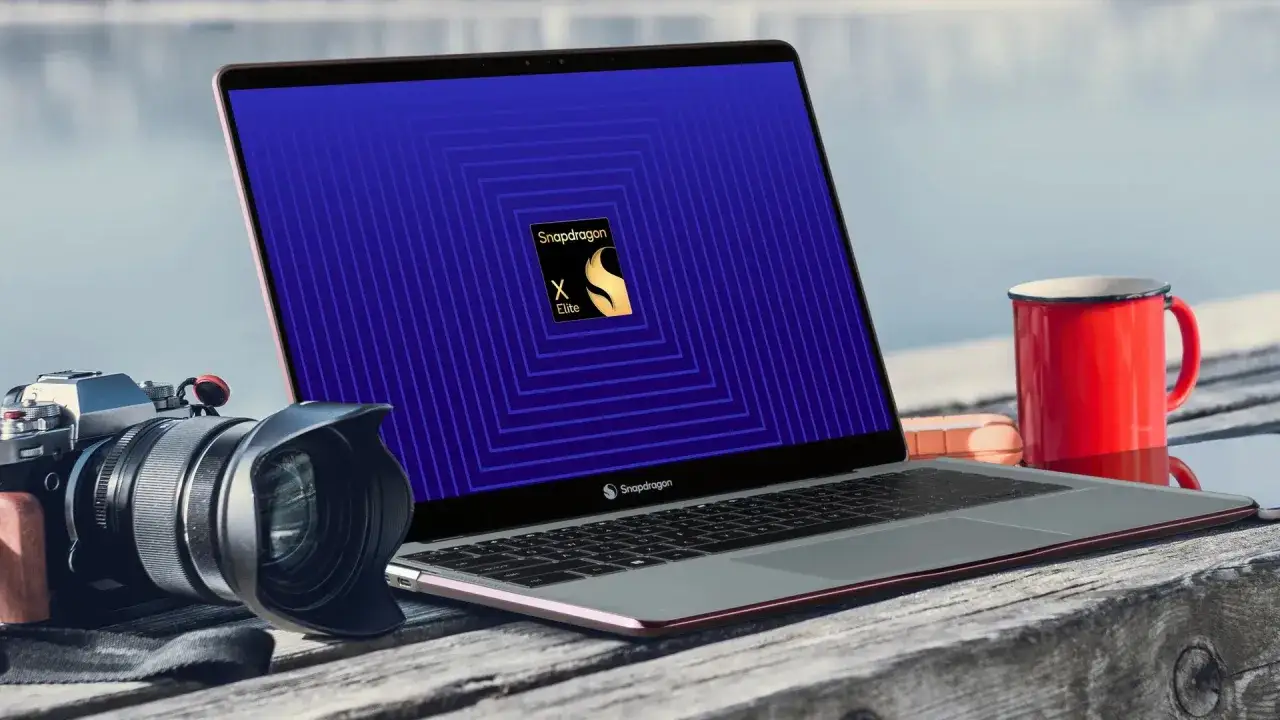 Laptop z logo Snapdragon X Elite na ekranie, obok aparat fotograficzny i czerwony kubek. Idealny zestaw dla twórcy mobilnego.