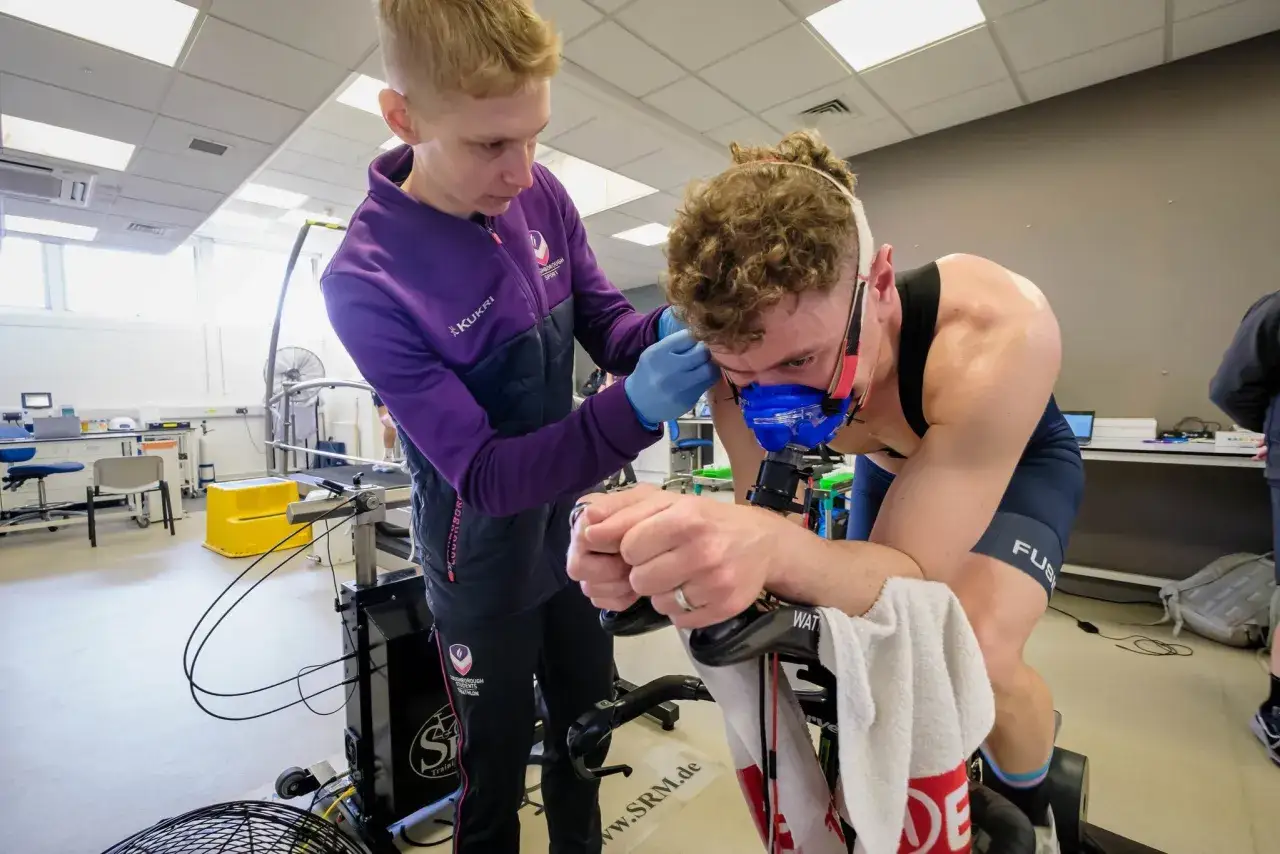 Sportler mit Maske misst seinen v2o max auf einem Ergometer, während ein Trainer die Messung vorbereitet.