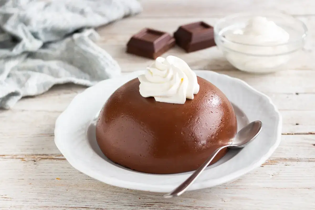 Un delizioso budino al cioccolato, guarnito con panna montata, su un piatto bianco. Accanto, due quadratini di cioccolato e una ciotolina di panna.