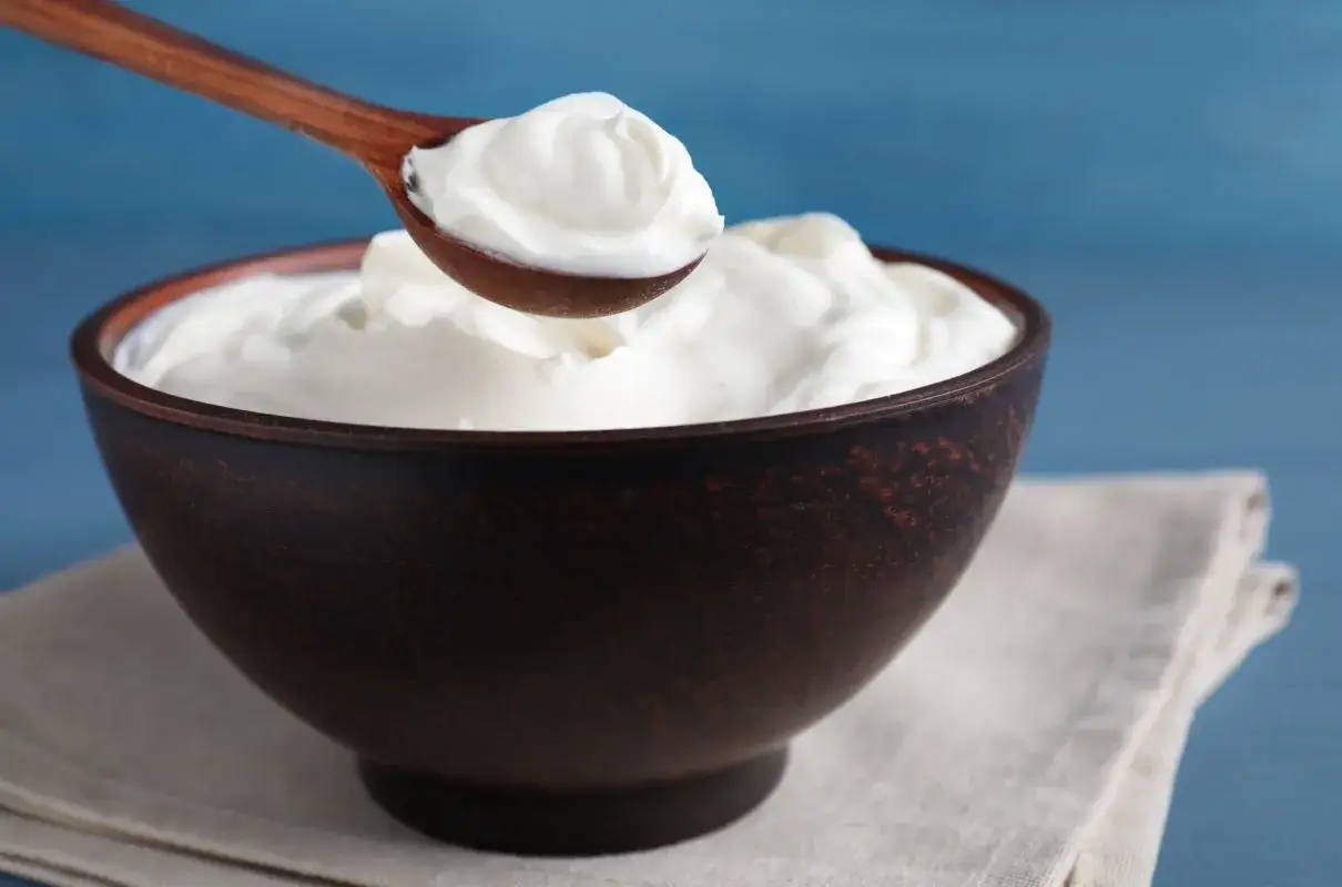 Gęsta, biała śmietana na drewnianej łyżce nad brązową miską. To właśnie creme fraiche, co to jest?