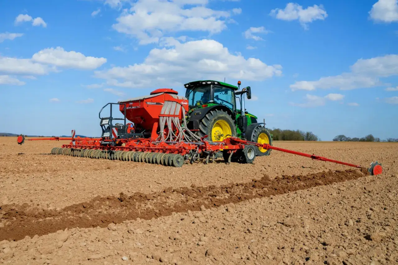 Tracteur vert et semoir rouge en action pour un semis direct sur un champ labouré sous un ciel bleu et nuageux.